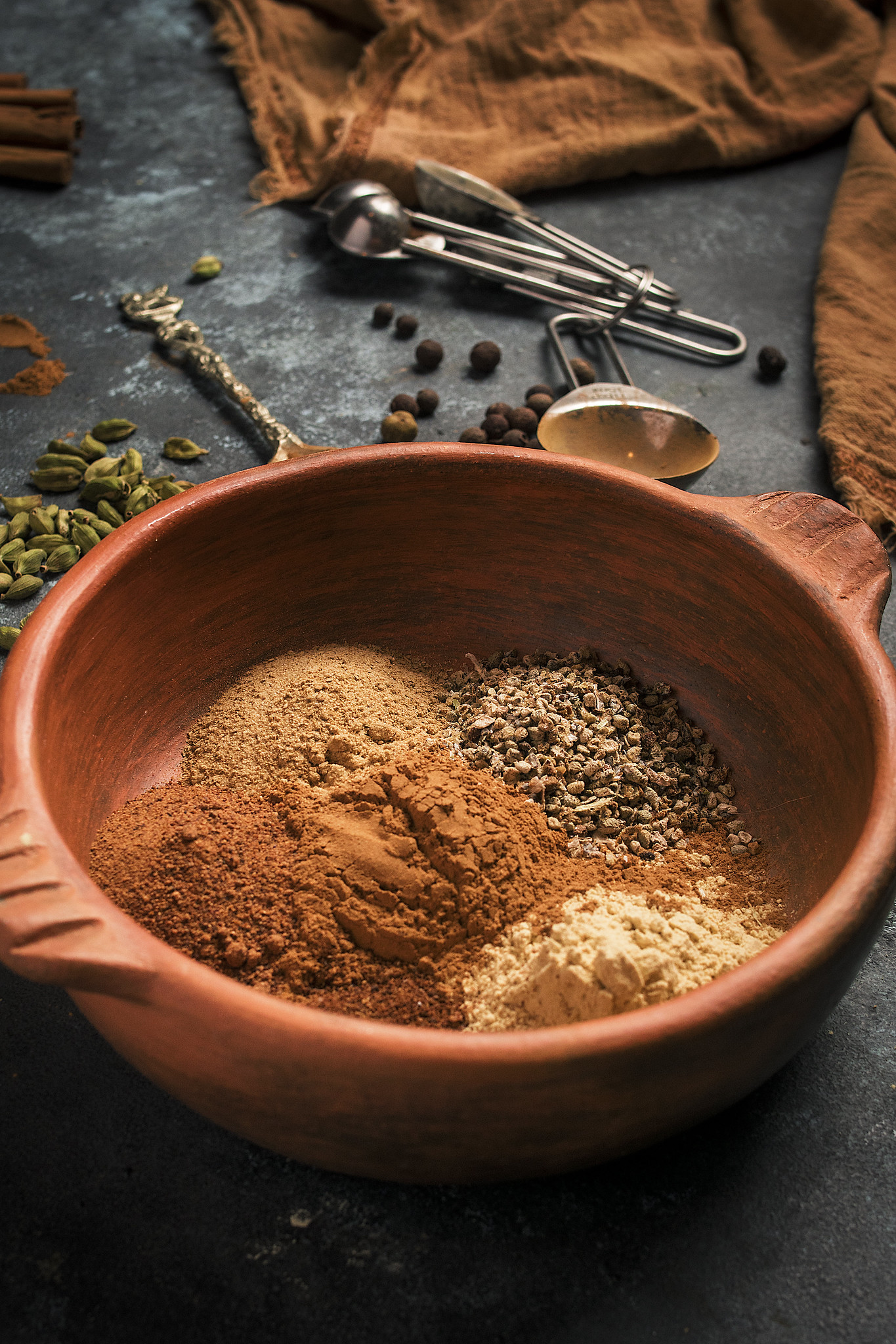 Ground spices measured into a terracotta bowl before mixing, with cardamom pods and measuring spoons nearby.